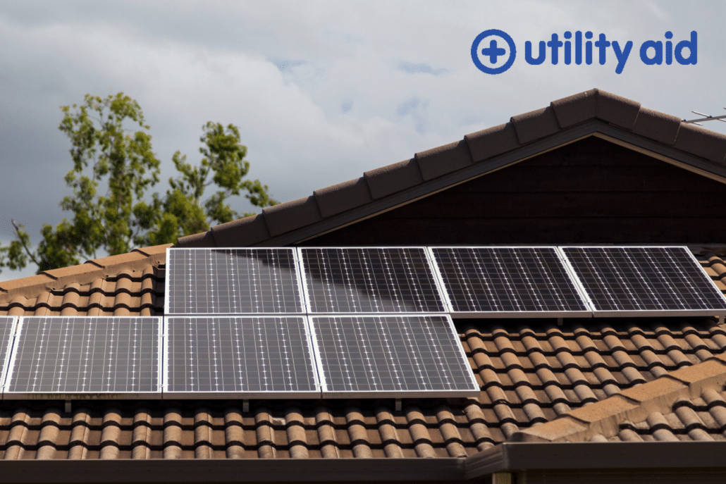 Solar panels installed on a tiled house roof under a partly cloudy sky, with the Utility Aid logo in blue at the top right corner. Trees are visible in the background.