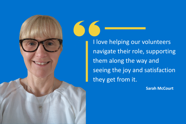 Graphic with a blue background featuring a person wearing a white top and a necklace. To the right, there is a large yellow quotation mark and a quote in white text that reads: “I love helping our volunteers navigate their role, supporting them along the way and seeing the joy and satisfaction they get from it.” Below the quote, the name Sarah McCourt is displayed in smaller white text.