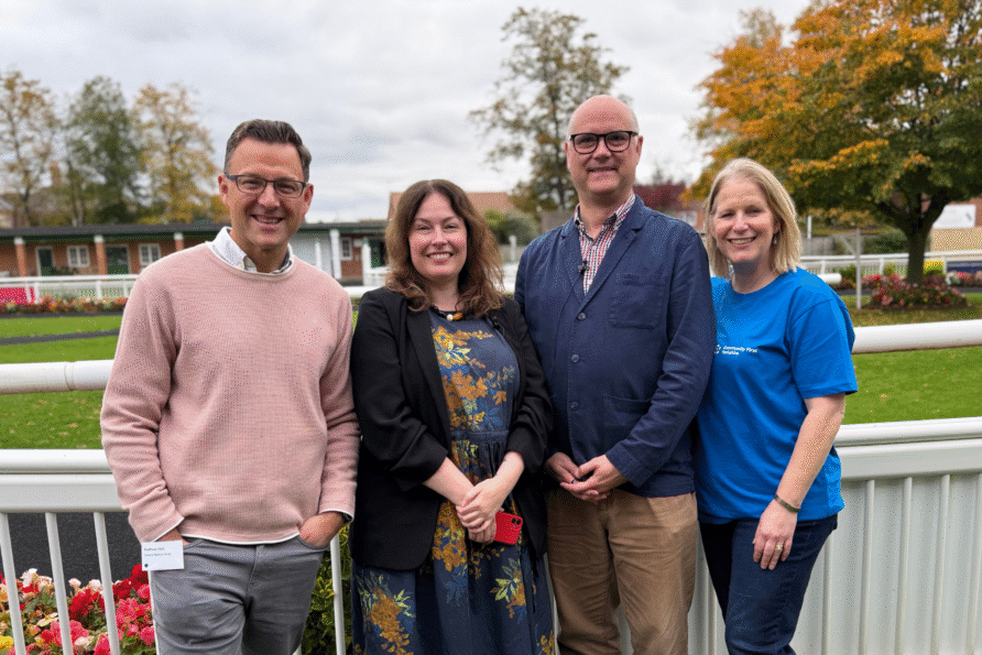 Four people standing outdoors in front of a white fence with a racetrack and flowerbeds behind them. The group is dressed in casual to smart-casual clothing, and the background shows green grass, colorful flowers, and trees with autumn foliage under a cloudy sky.
