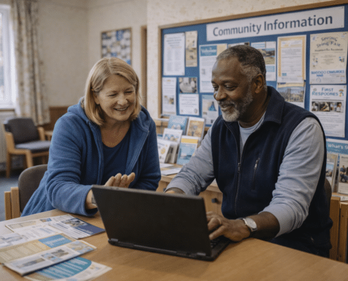 Two smiling adults sit at a table and look at a laptop in a community centre. Leaflets are spread out on the table, and a noticeboard labelled Community Information is visible in the background.