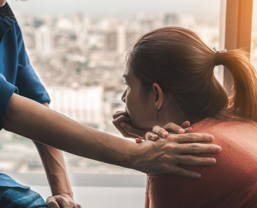 A person comforts a woman by gently placing a hand on her shoulder as she looks thoughtfully out of a window at a blurry cityscape, reflecting on her mental health challenges and the need for better mental health service.