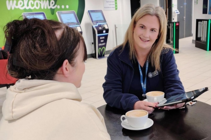 Two women sit at a table with coffee cups, one holding a tablet and smiling—a true trailblazer moment. Behind them are self-service machines and a green "welcome..." sign in a public indoor space.