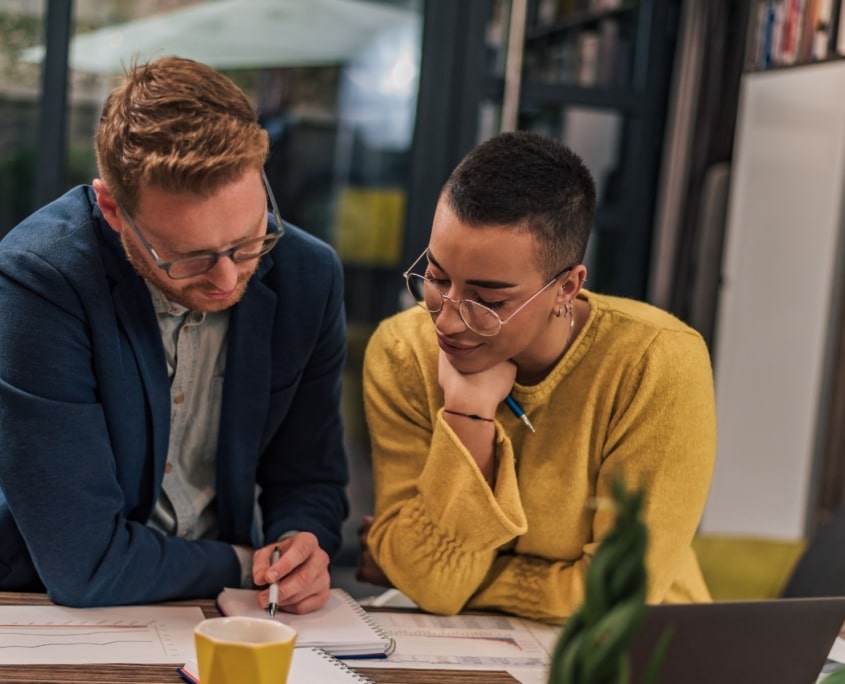 Two people sit at a table indoors, reviewing documents together. One is pointing at a notebook while discussing income sources and financial wellbeing, as the other listens attentively. A laptop, coffee cup, and papers are visible on the table.