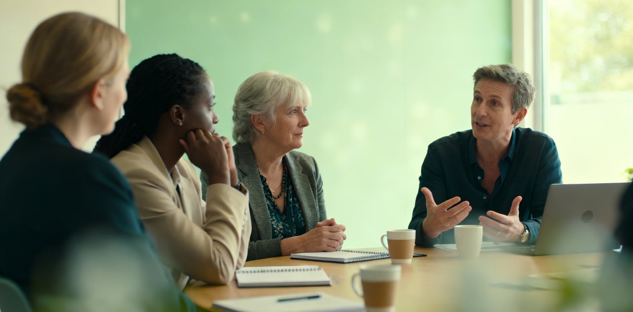 Four people sit around a table in a meeting room, engaged in discussion. Two women and one man listen as another man speaks, gesturing with his hands. Notebooks and coffee cups are on the table.