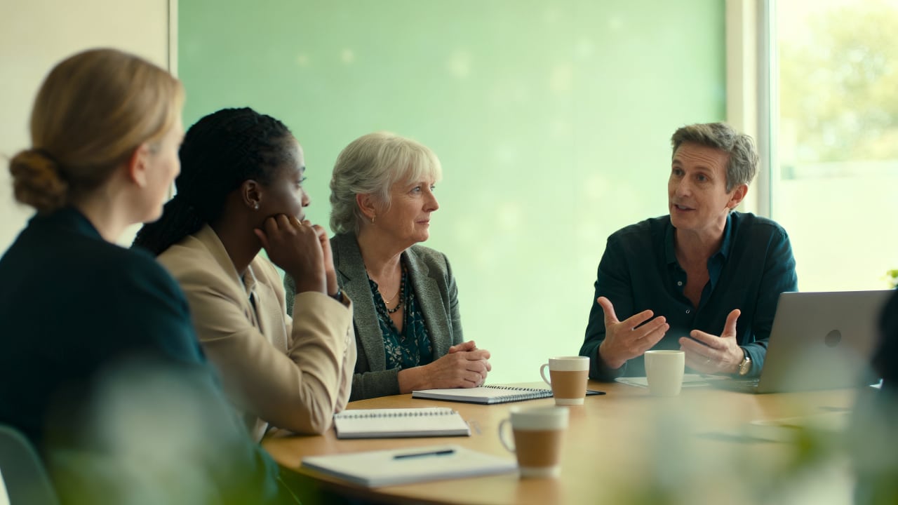 Four people sit around a table in a meeting room, engaged in discussion. Two women and one man listen as another man speaks, gesturing with his hands. Notebooks and coffee cups are on the table.