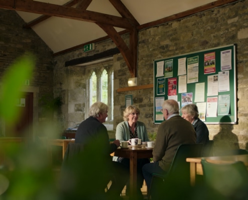 Four older adults sit around a table, chatting and smiling over drinks in a cosy, rustic room with stone walls, wooden beams, and a noticeboard with colourful notices in the background. Green plants are in the foreground.