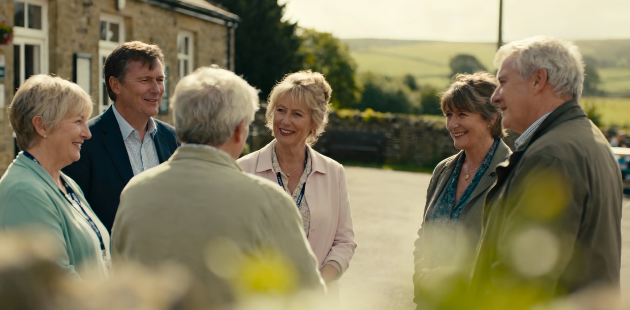 A group of six older adults, three men and three women, stand outside a stone building, smiling and chatting together on a sunny day with green hills in the background.