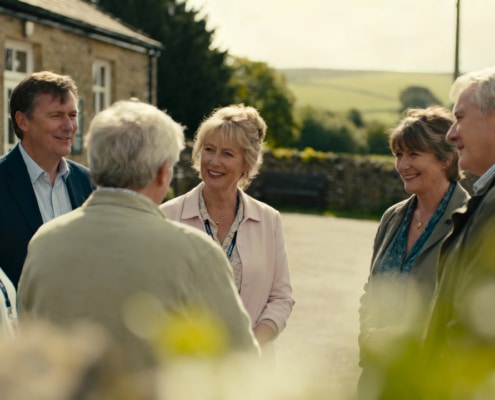 A group of six older adults, three men and three women, stand outside a stone building, smiling and chatting together on a sunny day with green hills in the background.