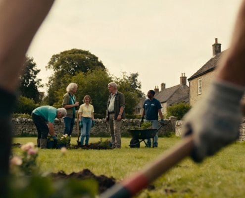 A group of people garden together on a grassy area in front of stone houses, with some planting flowers and others holding tools. The scene is framed by a person raking in the foreground.