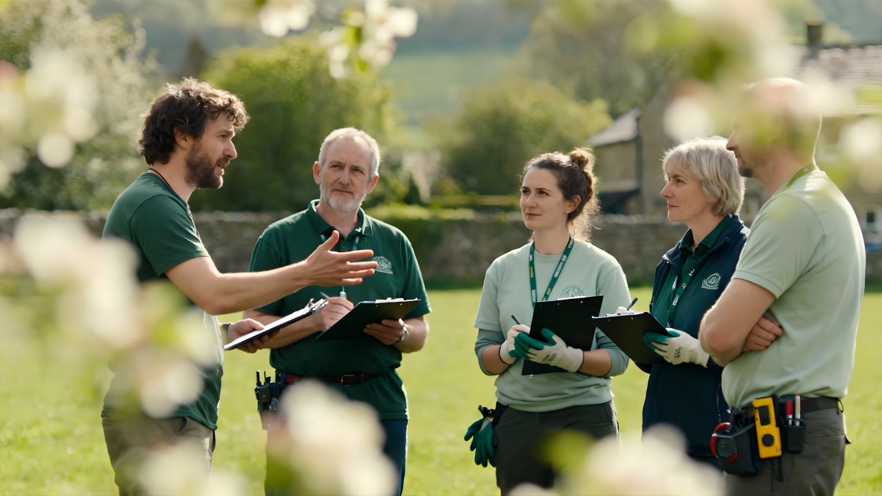 A man gestures while speaking to four people holding clipboards in an outdoor garden area. All wear green uniforms and appear to be having a discussion or training session. Flowering branches are visible in the foreground.