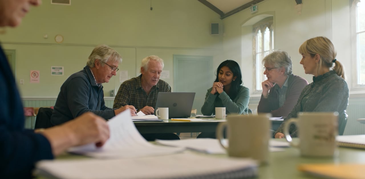 A group of six adults sits around a table in a bright room, engaged in discussion with papers, notebooks, and mugs in front of them. One man is using a laptop whilst others listen and take notes.