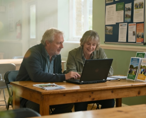 An older man and woman sit together at a wooden table, smiling while looking at a laptop. Informational leaflets and papers are on the table, with noticeboards and empty chairs in the background.