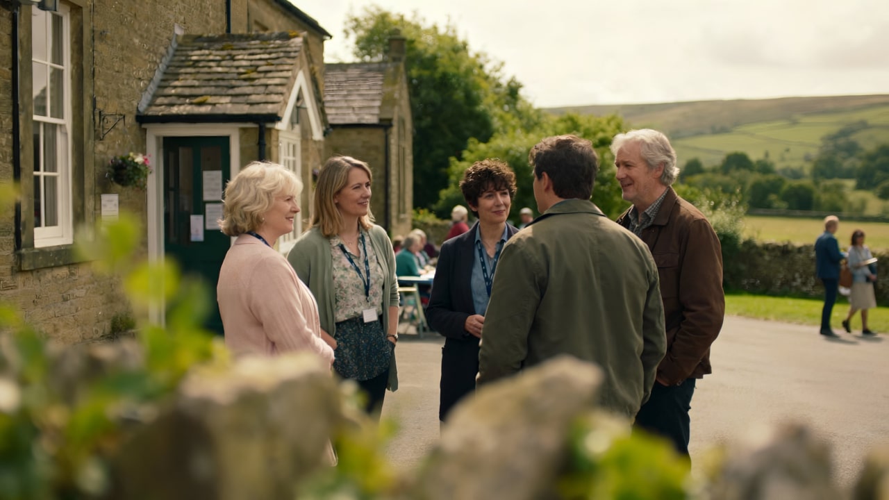 Five adults stand in a circle talking and smiling outside a stone building on a sunny day, with green hills and other people visible in the background. Some wear name badges, suggesting a social or community event.