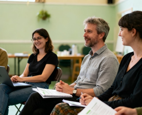 Five adults sit in a circle indoors, smiling and holding notebooks or a laptop, appearing engaged in a discussion or workshop in a bright, casual setting.
