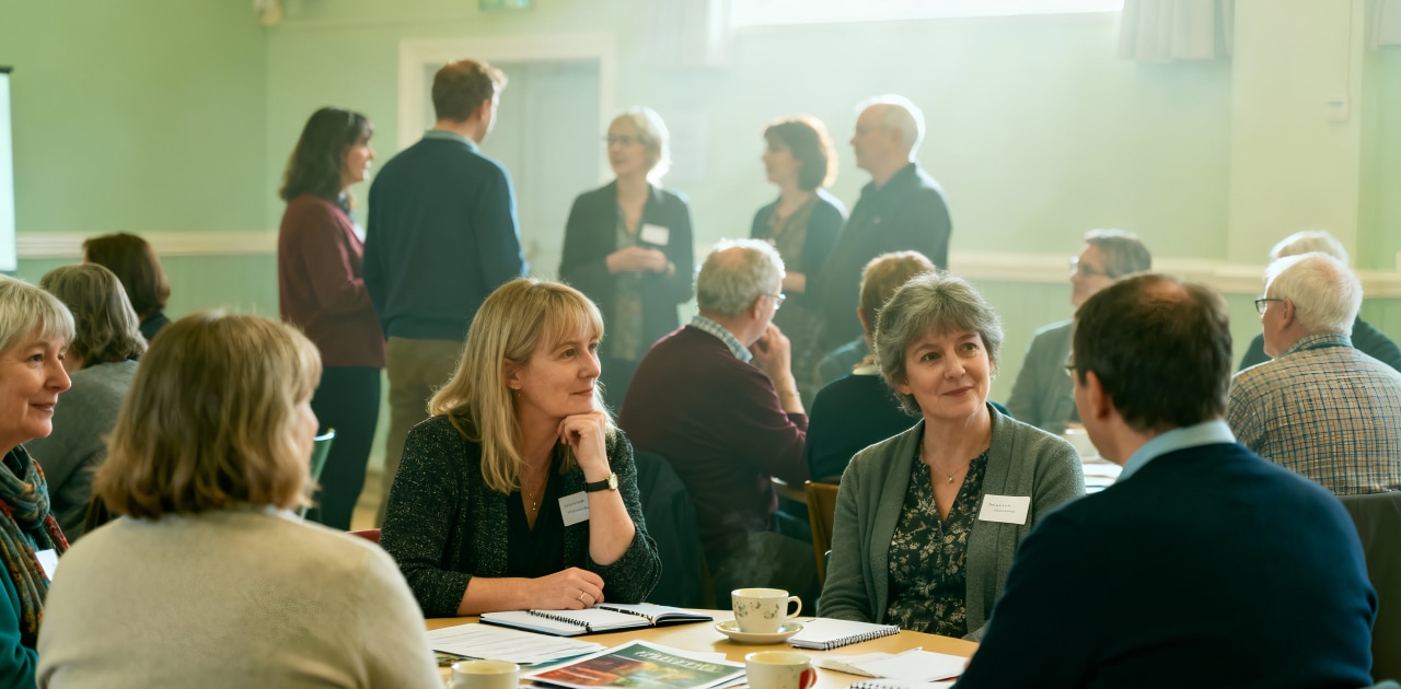 A group of adults sit at tables with notebooks and cups, engaged in discussion, whilst others stand and talk in the background in a bright, sunlit room, suggesting a community meeting or workshop.