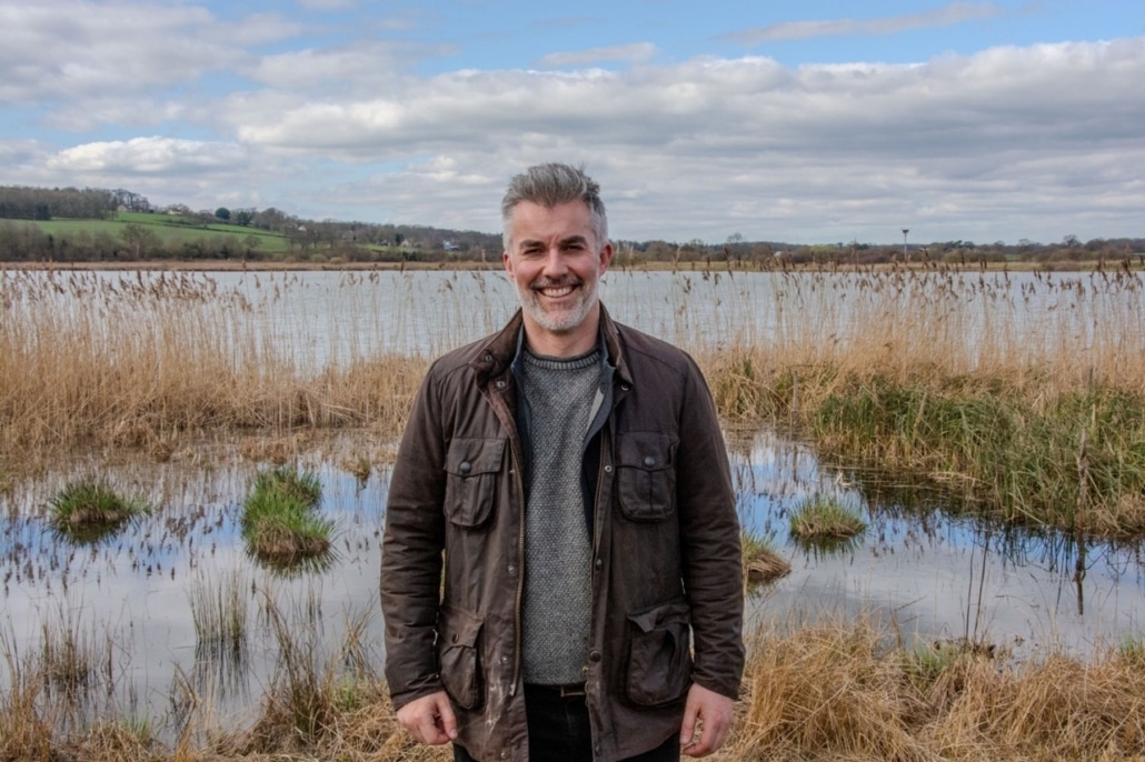 A man with grey hair and a beard stands smiling in front of a marshy lake, where community energy projects are bringing local power to life amid tall reeds, grassy patches, and distant green hills under a cloudy sky.