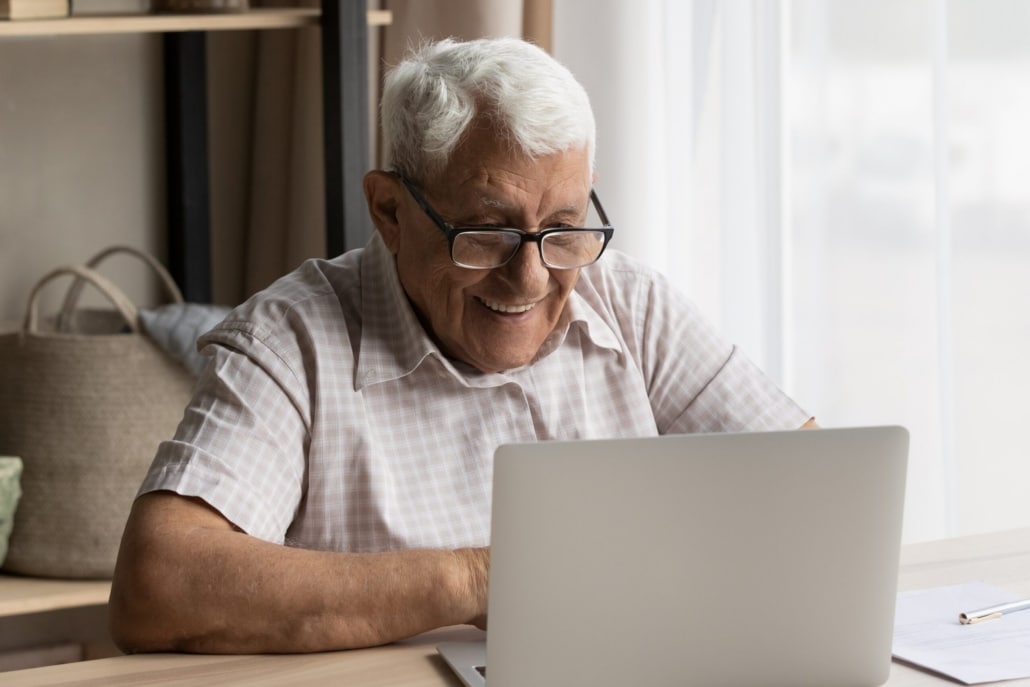 Smiling older man with glasses sits at a desk using a laptop, possibly completing an adult safeguarding survey, with sunlight coming through a window and shelves in the background.