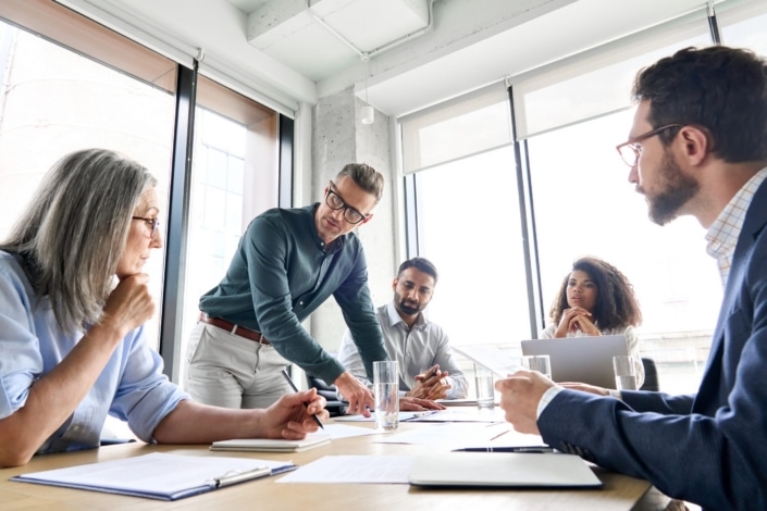 Five professionals sit around a conference table in a modern office, discussing Third Sector Trends among voluntary organisations in England and Wales. One man stands, gesturing to a document as the others listen. Natural light streams through large windows.