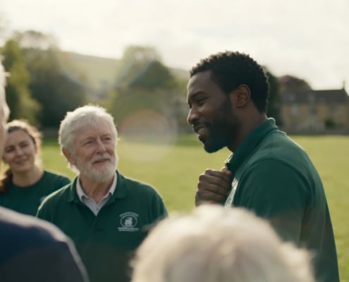 A group of people wearing green polo shirts stand and talk on a grassy field, with trees and houses in the background, enjoying a sunny day outdoors.