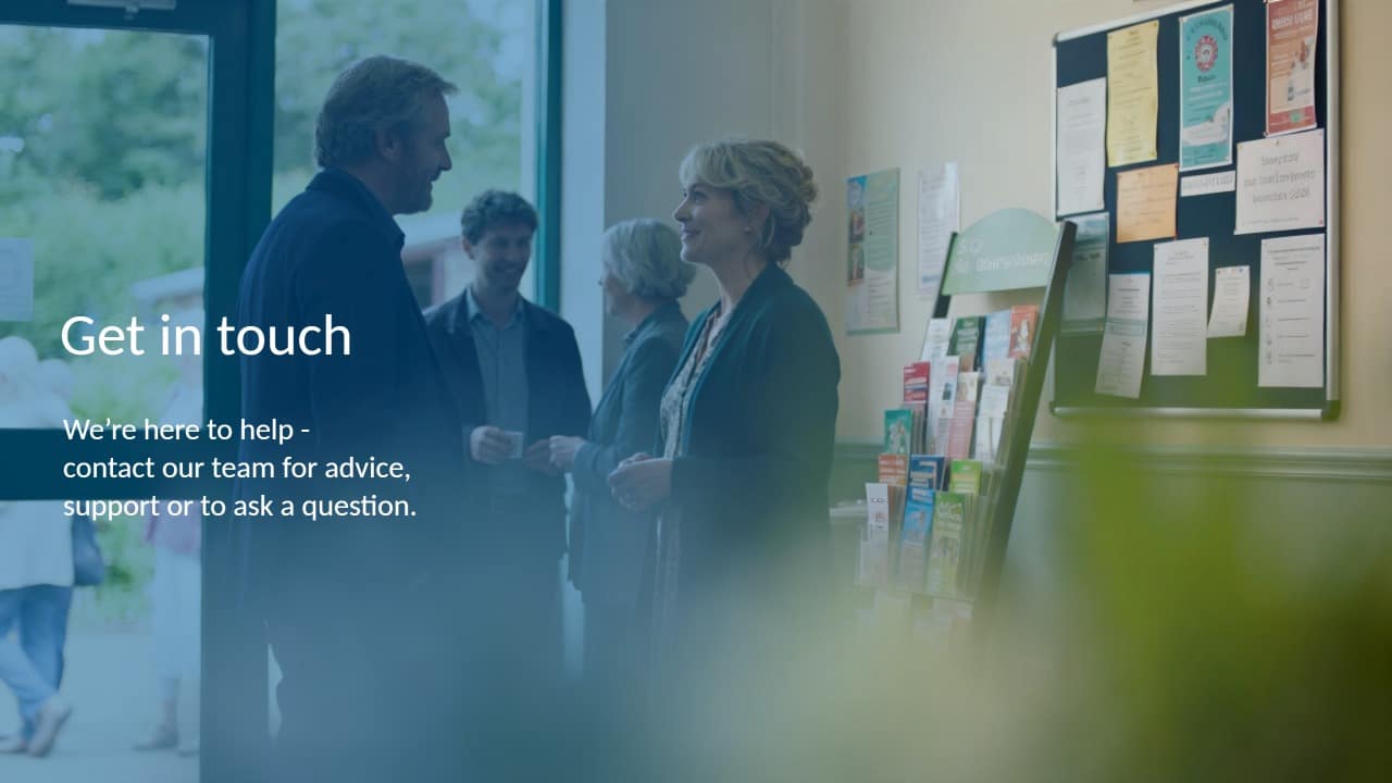 Two people stand and talk inside a community centre by a display of brochures. Other people are in the background. Text reads: Get in touch. We’re here to help—contact our team for advice, support or to ask a question.
