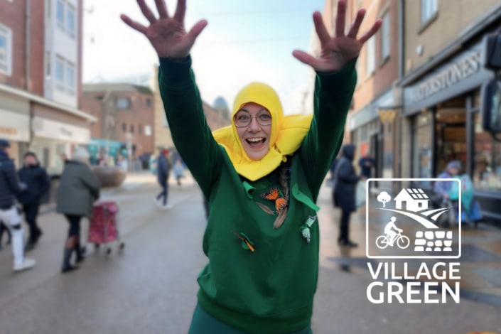 A person in a daffodil costume smiles with raised arms on a busy street during the Scarborough Pancake Race. The Village Green logo, featuring icons of a tree, house, bike and path, is overlaid on the right-hand side of the image.