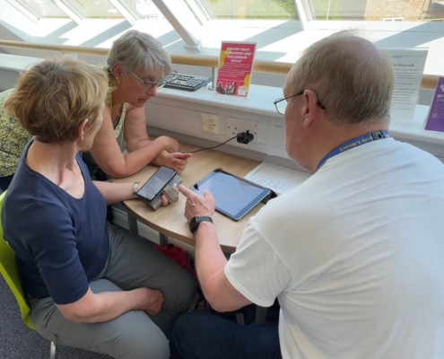 Three older adults sit at a table by a window, using a smartphone and tablet together. Connecting at their North Yorkshire Community Hub, they share smiles as bright daylight streams in.