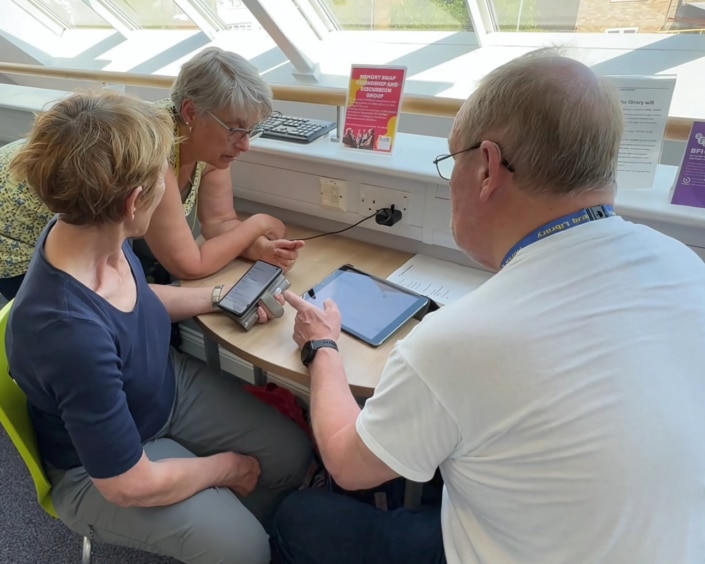 Three older adults sit at a table by a window, using a smartphone and tablet together. Connecting at their North Yorkshire Community Hub, they share smiles as bright daylight streams in.