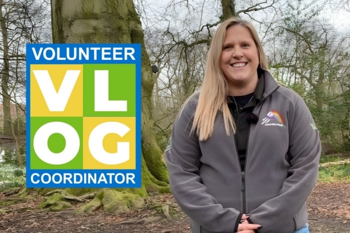 A woman in a grey jacket stands smiling in a wooded area in North Yorkshire. To her left is a colourful sign that reads Volunteer VLOG Coordinator in bold letters.