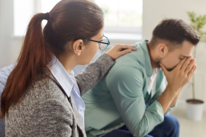 A woman with glasses comforts a distressed man who is sitting with his head in his hands. She rests her hand on his shoulder, supporting male victims of domestic abuse in a bright, indoor setting.