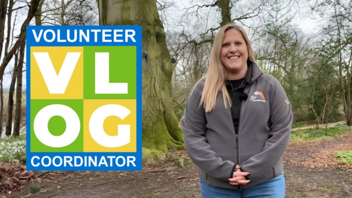 A smiling woman stands on a wooded path in North Yorkshire, wearing a grey jacket. To her left is a colourful sign that reads “VOLUNTEER VLOG COORDINATOR” in bold letters. Trees and greenery surround her, celebrating local volunteers.
