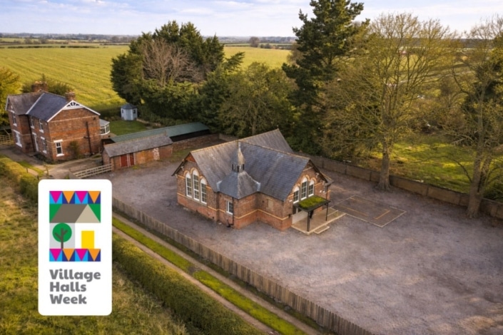 Aerial view of Sandhutton Village Hall with a slate roof, surrounded by trees and open fields. In the foreground, a Village Halls Week banner celebrates community and bringing people together with a colourful graphic of a hall and tree.
