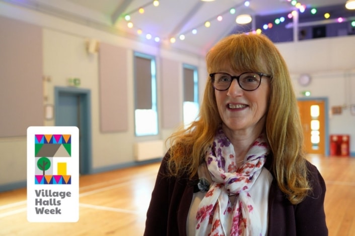 A smiling woman with glasses and a patterned scarf stands in a brightly lit village hall decorated with colourful fairy lights, celebrating Village Halls Week and showcasing how these spaces boost community wellbeing. The Village Halls Week logo appears on the left.