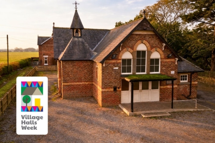 SandHutton Village Hall, a red brick building with arched windows and a small bell tower, sits among trees and fields. The Village Halls Week logo in the lower left corner highlights its role in supporting a greener future.