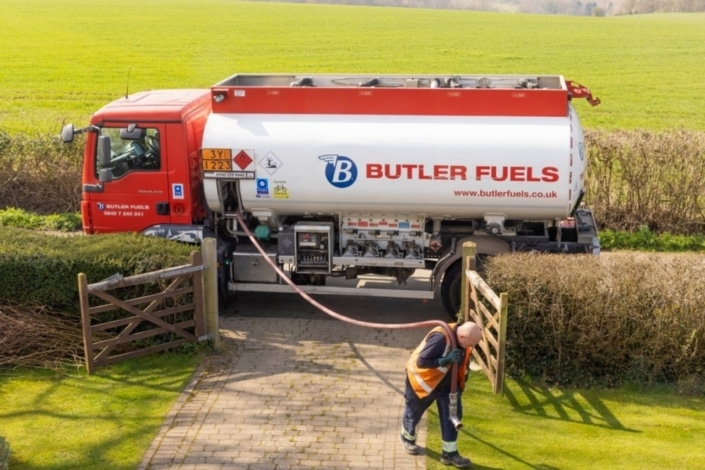 A fuel delivery lorry from Butler Fuels is parked outside a house, with a worker in a high-visibility vest handling a hose on a drive. The scene is set in a rural area with green fields in the background.