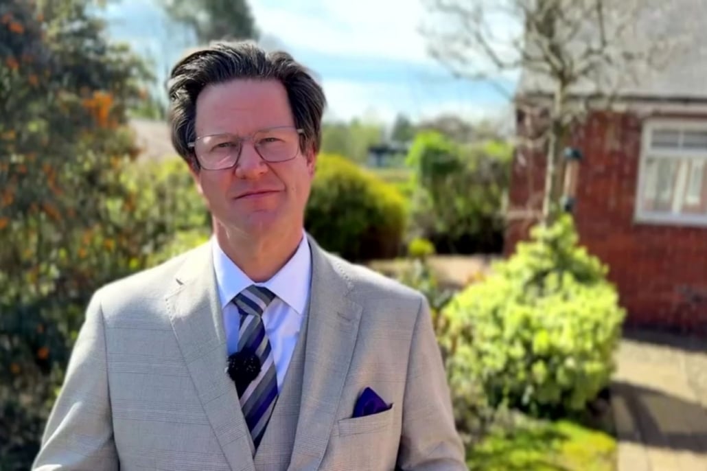 Alec Shelbrooke MP, in a light grey suit, striped tie, and glasses, stands outdoors in a sunny garden with green bushes and a red brick house, engaging with local communities.