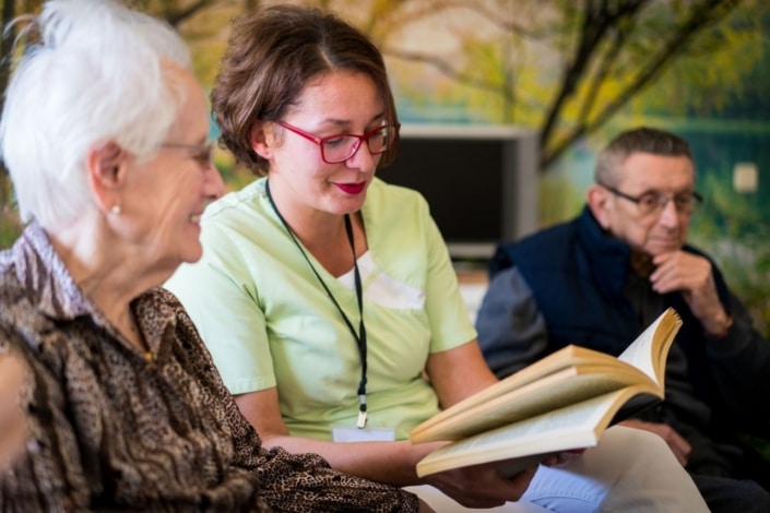 A carer sits between two older adults, holding and reading from a book. The group appears engaged, with a nature-themed mural in the background, highlighting the supportive spirit of the VCSE Workforce in North Yorkshire and Humber.
