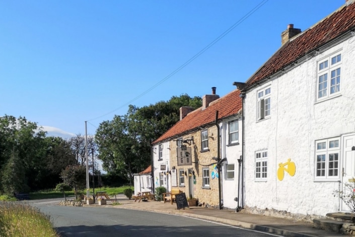 A quaint English village street with stone cottages, a red-tiled roof, and a B&B sign. Outdoor tables sit by the road, trees line the background under a clear blue sky—perfect for a rural housing conference near Ripon Cathedral.