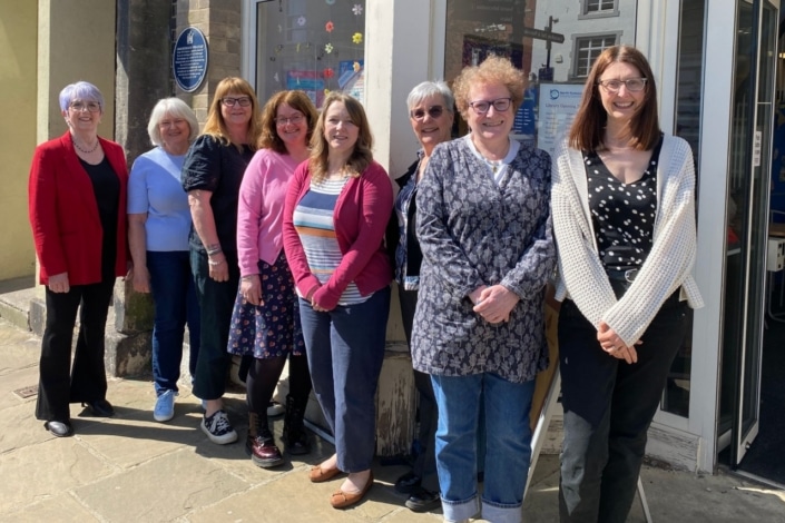 Eight women, Age Friendly Ambassadors, stand in a row outside a building on a sunny day, smiling at the camera. Casually dressed, they appear to be part of an age friendly team on a stone paved pavement.
