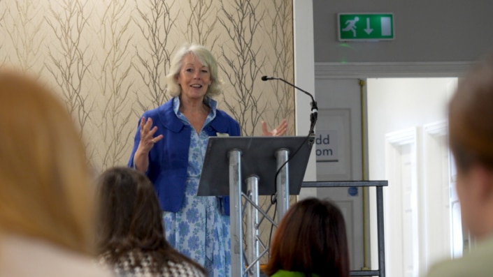 An older woman with grey hair, wearing a blue jacket and floral dress, speaks at a lectern during an inspiring morning event in front of an audience in a well-lit room.