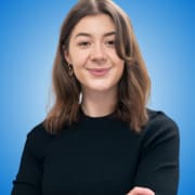 A young woman with shoulder-length brown hair and a slight smile stands confidently with arms crossed, embodying the poise of workshop hosts, wearing a black top against a bright blue gradient background.