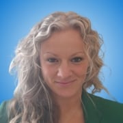Portrait of a woman with wavy, light blonde hair, wearing a green blazer and necklace, smiling softly. The background is a gradient of light to medium blue—an ideal look for workshop hosts or speakers at professional events.