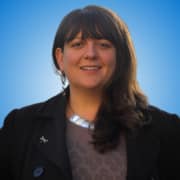 A woman with long brown hair and a fringe, wearing a black coat, a patterned top, a large necklace, and earrings, smiles in front of a blue gradient background—an inspiring presence among fellow speakers.