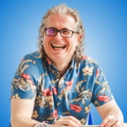 A man with curly grey hair and glasses, likely one of the workshop hosts or panellists, smiles broadly while wearing a blue floral shirt and jewellery, sitting at a table with a blue gradient background.