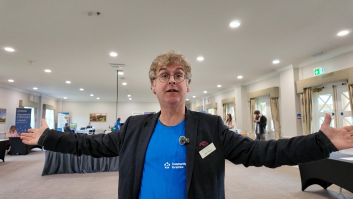 A person with curly hair, glasses, and a blue shirt stands indoors with arms outstretched, radiating energy. The room’s tables, banners, and people hint at an event or conference like Create 26 brimming with fresh ideas.
