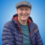 A smiling man wearing glasses, a grey flat cap, a blue quilted jacket, and a teal shirt sits in front of a bright blue gradient background—perfectly capturing the approachable style often seen among inspiring speakers and workshop hosts.
