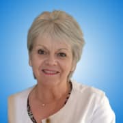 Smiling older woman with short grey hair, white top, and necklace, posing in front of a blue gradient background—ideal for profiles of inspiring speakers or distinguished panellists.