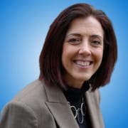 A woman with shoulder-length brown hair wearing a tan blazer, black top, and a chunky silver necklace smiles at the camera against a blue gradient background—perfect for introducing workshop hosts or speakers.