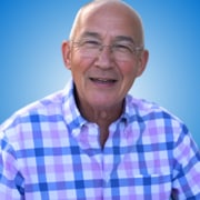 Smiling older man with glasses and a bald head, wearing a blue, purple, and white checked shirt, stands against a blue gradient background—perfect for workshop hosts or speakers at professional events.