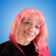 Woman with long pink hair, wearing hoop earrings and a black top, smiling in front of a bright blue background—perfect for event speakers or workshop hosts.