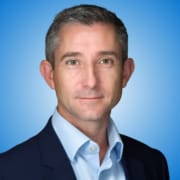 A man with short grey hair, wearing a dark suit jacket and light blue shirt, looks at the camera with a neutral expression. Set against a blue gradient background, he is among renowned speakers and workshop hosts in his field.